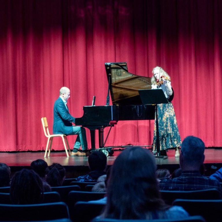 A man playing piano on a stage with red curtain backdrop and woman standing and playing the violin.