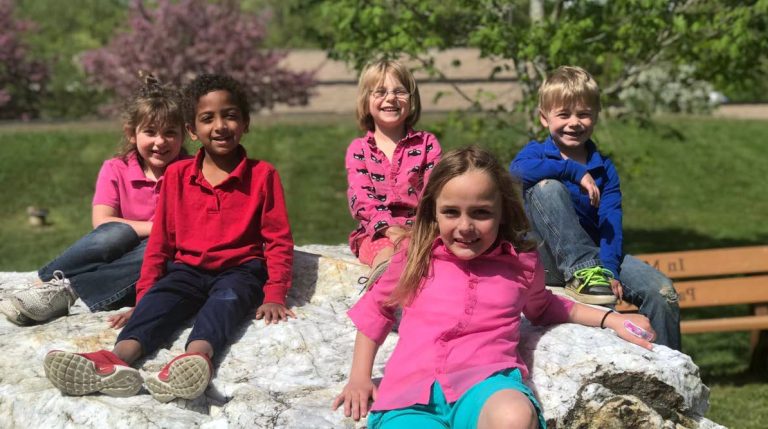A group of five children sitting on a large rock and smiling,
